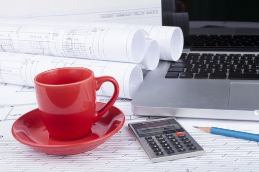 Architect blueprints with a calculator and a silver laptop with blue pencil and red cup of coffee