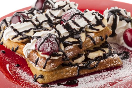 Belgian waffle with chocolate syrup whipped cream and banan slice with red cherry on red plate and sugar powder snow
