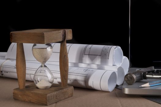 Architectural plans rolled with silver laptop hammer and a pencil and old wood hourglass