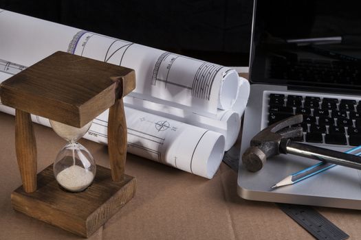 Architectural plans rolled with silver laptop hammer and a pencil and old wood hourglass