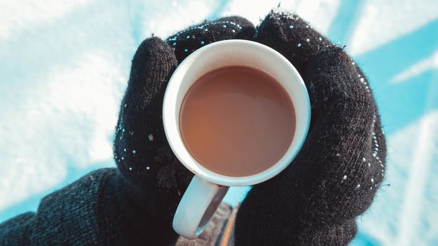 Warm cup of hot coffee warming in the hands in mittens. Womens hands on the background of snow, winter