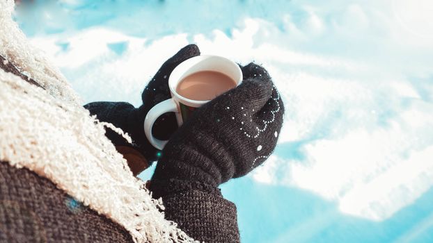 Warm cup of hot coffee warming in the hands in mittens. Womens hands on the background of snow, winter