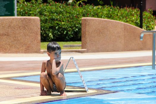 The little frustrated boy sits in swimming trunks near the swimming pool on a sunny summer day.