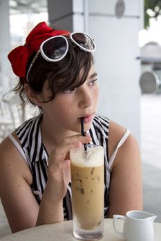A teenage girl with pretty freckles on her face, dressed in a summer blouse and with sun glasses on her head, drinks ice coffee through a straw in a cafe near the window.
