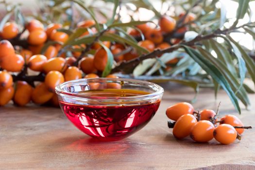 Sea buckthorn oil in a glass bowl on a wooden table, with sea buckthorn berries and leaves in the background