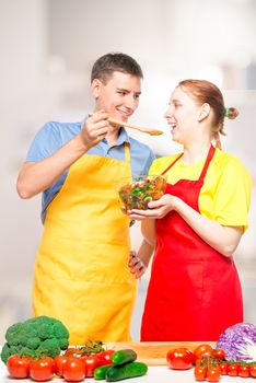 a man with a wooden spoon and a girl with a bowl of salad try the food cooked together in the kitchen