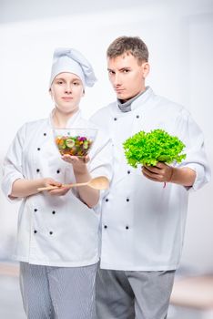 young cooks in suits with salad in hands on gray background