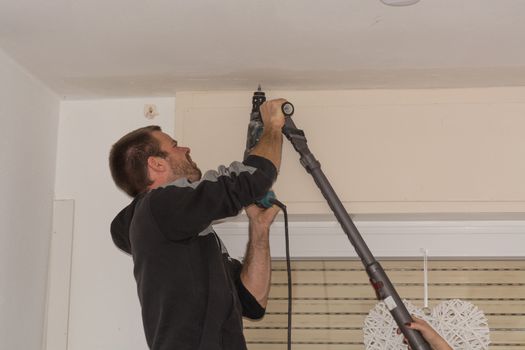 The man drills a hole in the ceiling before mounting a curtain rod