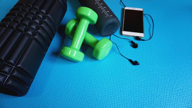 Foam Roller with green dumbbells and water bottle and telephone with headphones - Gym Fitness Equipment Blue background self Myofascial Release - MFR.