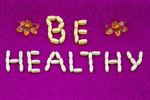 The inscription Be healthy, created from pills and capsules with vitamins and dietary supplements on a pink monophonic background. Copy space.
