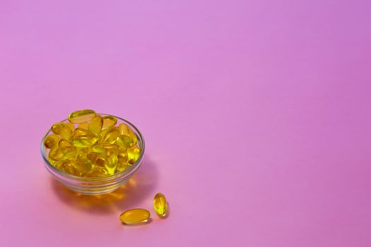 Yellow transparent capsules with fish oil containing Omega-3 acids in a transparent cup on a pink background.