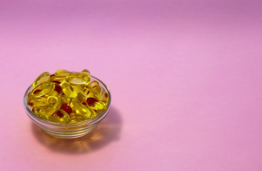 Yellow transparent capsules with fish oil containing Omega-3 acids in a transparent cup on a pink background.