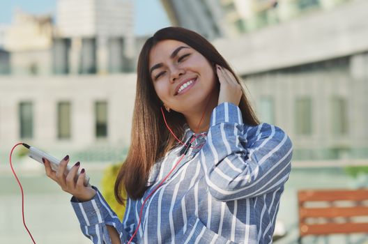 Portrait of a cheerful girl who walks around the city and listens to music from a mobile phone in vacuum headphones, raising her head up and closing her eyes