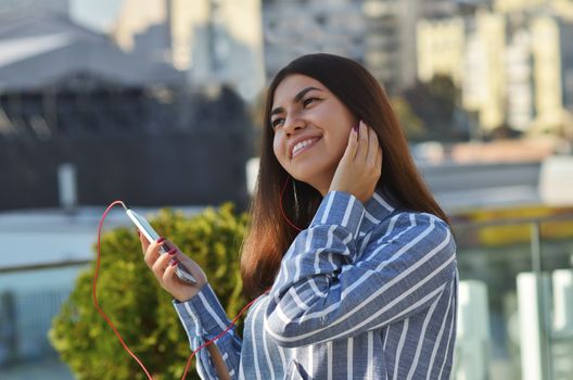 Portrait of a cheerful girl who walks around the city and listening to music from a mobile phone in vacuum headphones