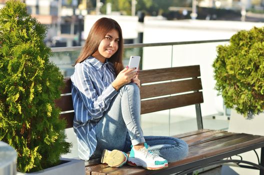 A modern girl in a beautiful shirt and sneakers uses the phone and sits on a bench in the city Park