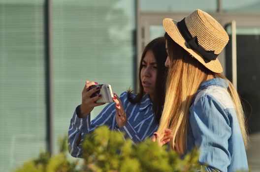Two young girls communicate in social networks by phone walking on the street on a Sunny day