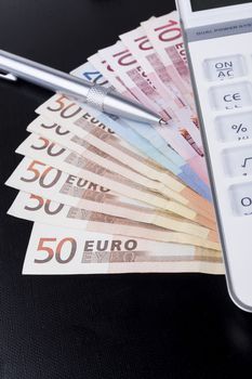 White calculator on top of euro banknotes on black background with silver pen