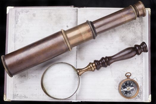Old telescope with magnifing glass and a compass on an old notebook with black background