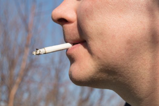 Young handsome man holding a cigarette in his mouth. Stands against the blue sky.