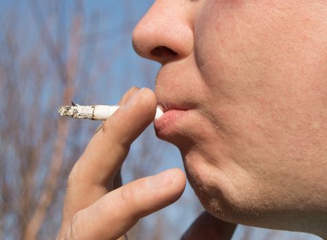 Young handsome man holding a cigarette in his mouth. Stands against the blue sky.