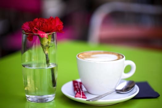 A white cup on a saucer with hot coffee on a green table in a street cafe, next to it is a glass with fresh carnation. Macro photo, blurred background.