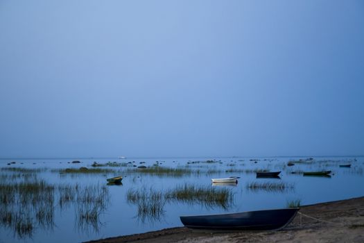 Fishing boats are anchored in a row, reflected in a clear lake at dusk.