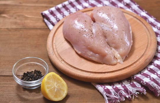 Raw chicken breast sprinkled with black pepper and salt lies on a round wooden Board, next to a lemon. Horizontal close-up photo