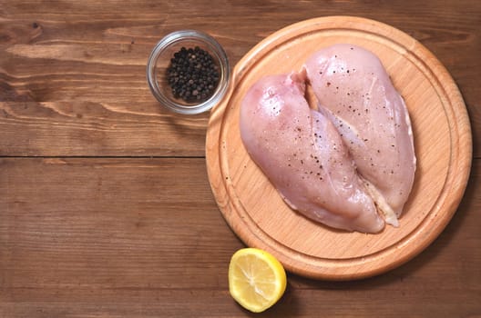 Two raw chicken fillets lie to the right in the shape of a heart on a wooden Board against a tree background. The meat is located in the center, next to a lemon with black pepper