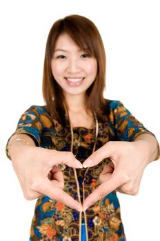 Southeast Asian girl wearing batik kebaya with hand forming a heart shape, standing isolated on white background.