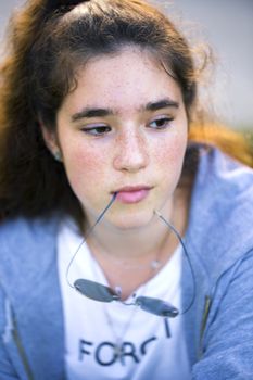 A portrait of a sad, pensive teenager girl with freckles and long brown hair who holds in her mouth a bow of glasses and sadly looked down.