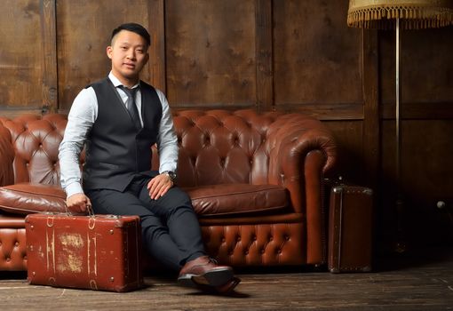 A young man in a suit sits on a leather brown sofa with a suitcase