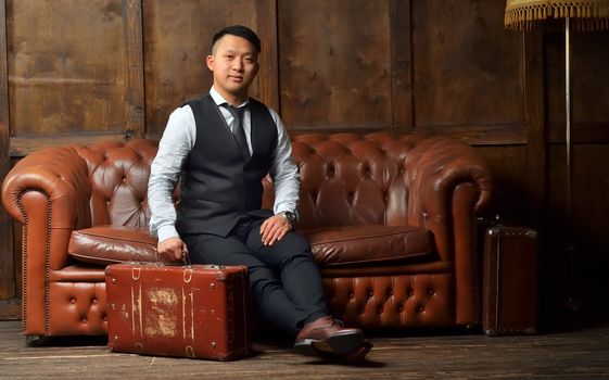 A young man in a suit sits on a leather brown sofa with a suitcase