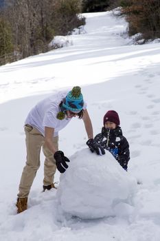 A young guy in a T-shirt and a cap makes a snowman with his younger brother in the winter forest on a nice sunny day.