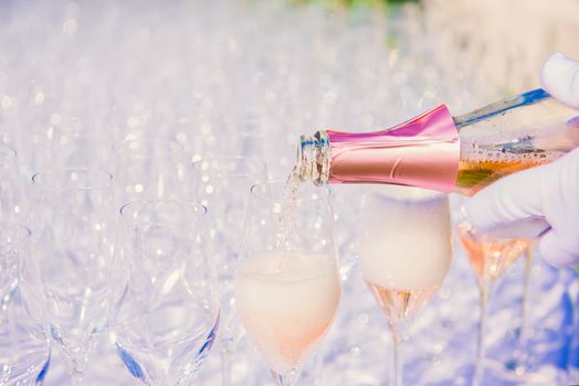 Waiter pouring champagne, sparkling wine into glasses at outdoor reception, wearing white gloves, wine foaming in glass, nobody, copyspace