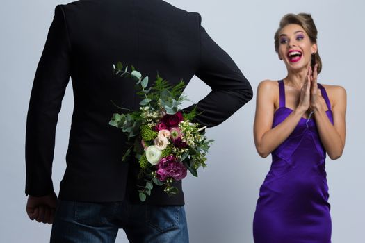 Man brings flowers to woman and hiding them behind back, womanin evening dress is surprised and happy