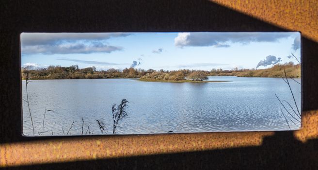 view from a viewpoint at water in Holland with a pond, water plants small islands and the smoke from the europoort industry at the background