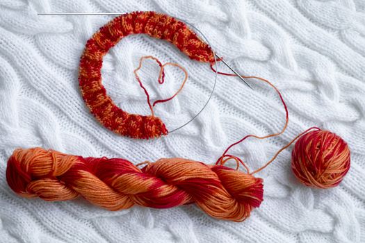 Knitting project in progress. A piece of knitting with a ball and a skein of red-orange color and knitting needles against a white knitted plaid.