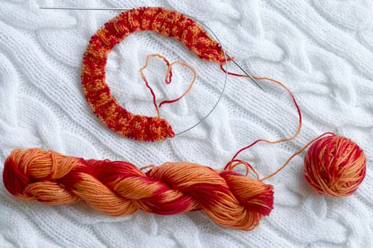 Knitting project in progress. A piece of knitting with a ball and a skein of red-orange color of sectional dyeing and knitting on the background of a white knitted plaid.