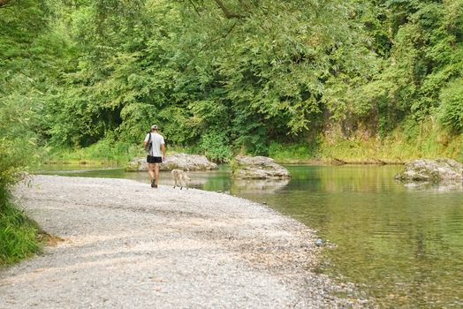 A young man with a cap walks by the river with his dog, sunny day