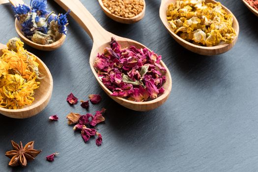 Herbs on wooden spoons on a dark background with copy space. Dried calendula, cornflower, rose petals, fenugreek seeds, mullein, star anise.