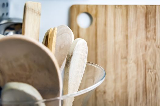 Wooden kitchen utensils in a glass container with a wooden cutting board in the background. Kitchen interior shot. Home decor and cooking concept