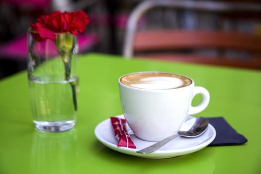 A white cup on a saucer with hot coffee on a green table in a street cafe, next to it is a glass with fresh carnation. Macro photo, blurred background.