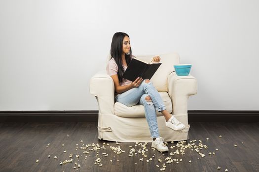 young woman sitting on couch, reading a book and eating popcorn while leaving a mess