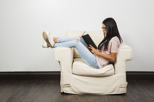 Twenty something woman sitting on the arm of a couch, with her feet up, reading a book