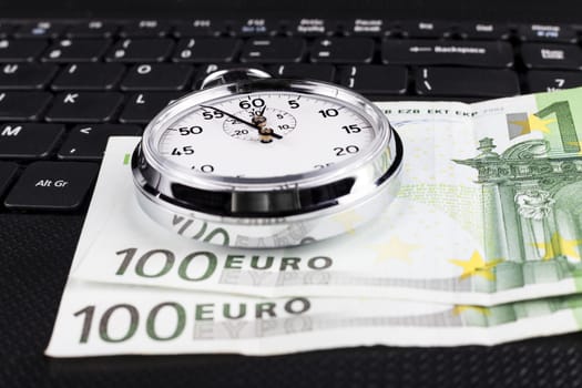 Silver stopwatch on black laptop keyboard with 100 euro bills