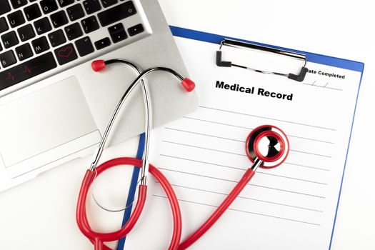 Red stethoscope on  blue clipboard  with medical record close-up with silver colored laptop