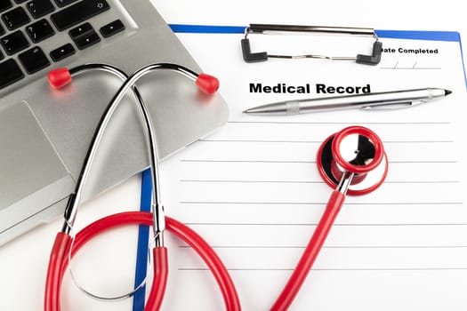 Red stethoscope on  blue clipboard  with medical record close-up with silver colored laptop