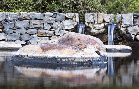 big rock in  a water pool with reflection an two small waterfalls and some vegetation