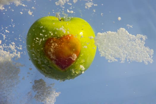 Green apple with a red heart on its side, surrounded by snow against a blue sky