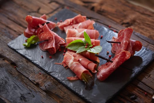 Slices of Italian prosciutto crudo or jamon with fresh basil leaves on a black background. Mediterranean appetizer or antipasto. Raw ham. Top view. Copy space.